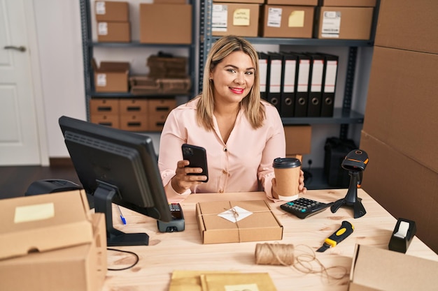 Une jeune femme hispanique travaillant dans un e-commerce utilise un smartphone et boit du café au bureau.