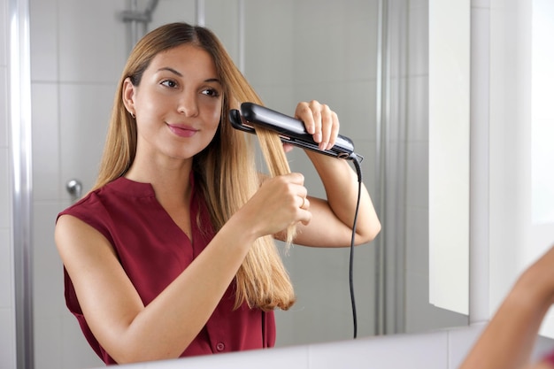 Photo une jeune femme hispanique utilise un redresseur à vapeur pour coiffer ses cheveux dans le miroir de la salle de bain.