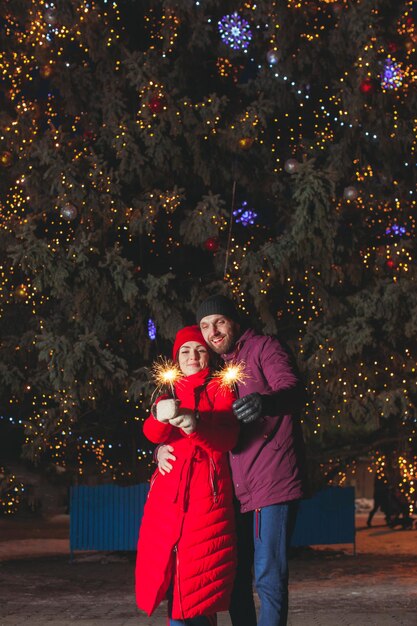 Jeune femme et homme amoureux, debout étreignant près d'un grand sapin, tenant des lumières scintillantes dans leurs mains. Portrait en plein air d'un couple élégant en vêtements d'hiver chauds