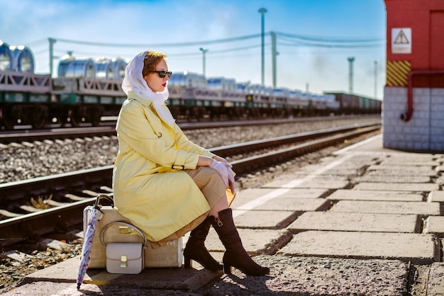 Une jeune femme en imperméable jaune et lunettes noires est assise sur une valise sur le quai d'une gare ferroviaire