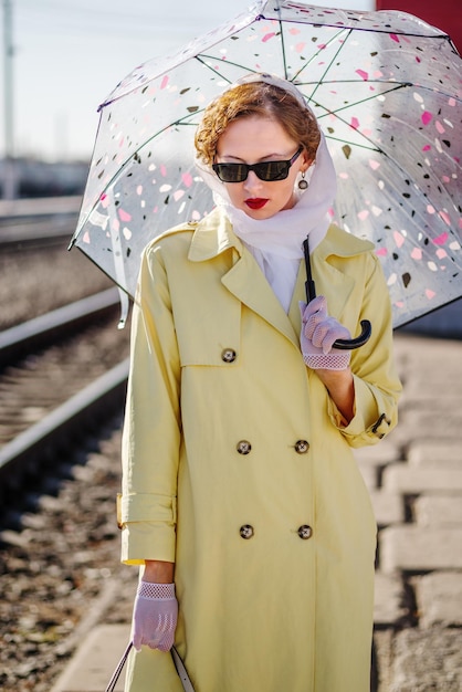 Une jeune femme en imperméable jaune et lunettes noires avec parapluie dans ses mains Portrait de style rétro