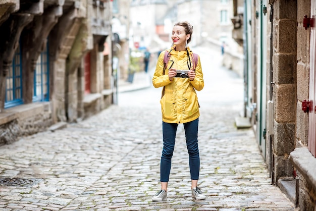 Jeune femme en imperméable jaune marchant avec sac à dos et appareil photo dans le village de Dinan en région Bretagne en France