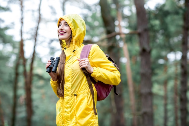 Jeune femme en imperméable jaune randonnée avec jumelles et sac à dos dans la pinède verte. Observation des oiseaux dans la forêt