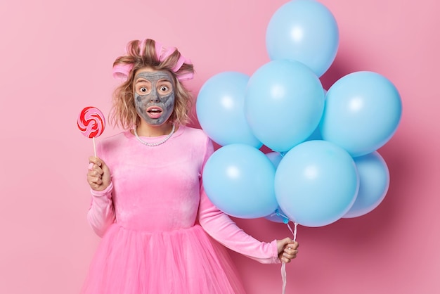 Une jeune femme impressionnée et choquée regarde les yeux sur écoute, applique un rouleau à cheveux et un masque de boue contient des bonbons sucrés sur un bâton et un tas de ballons gonflés se prépare pour des vacances isolées sur fond rose