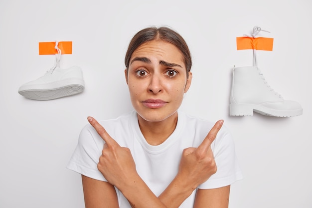 Une jeune femme inconsciente et perplexe pointe de côté la basket et la botte hésite à choisir quoi porter vêtue d'un t-shirt décontracté isolé sur un mur blanc