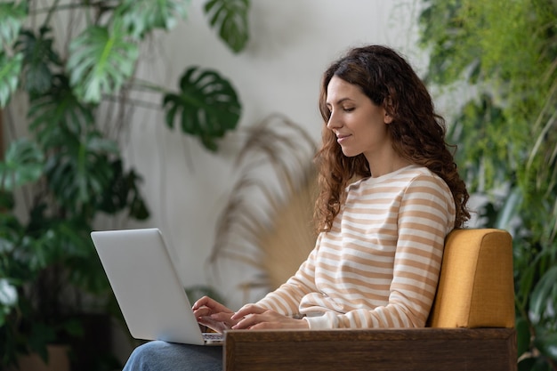 Jeune femme indépendante assise dans un fauteuil en serre travaillant sur un ordinateur portable entouré de plantes d'intérieur