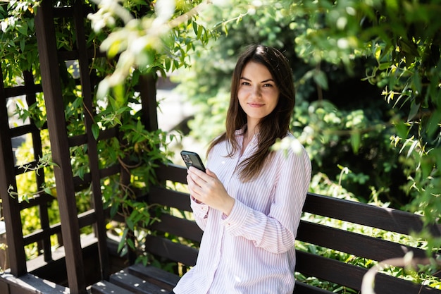 Jeune femme indépendante en chemise s'asseoir sur un banc dans le parc d'été à l'extérieur se reposer et utiliser un téléphone portable