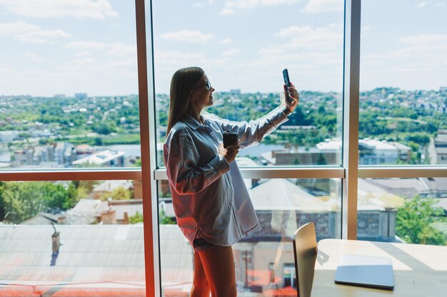 Une jeune femme indépendante à lunettes se tient avec un café et parle au téléphone près d'une grande fenêtre et regarde la ville Une fille en chemise et short se tient dans un café près de la fenêtre