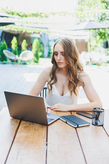 Jeune femme indépendante positive assise à une table avec un ordinateur portable et travaillant sur une terrasse verte regardant l'écran et travaillant sur un projet à distance dans un café