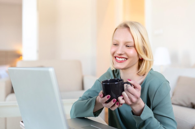 Jeune femme indépendante positive regardant une vidéo sur un blog sur netbook et prenant un café dans une maison confortable Belle jeune femme d'affaires travaillant sur un ordinateur portableFreelancer se connectant à internet