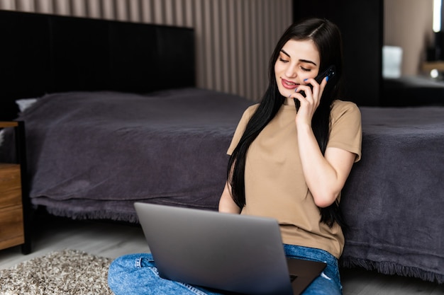 Jeune femme indépendante travaillant à domicile, assise sur le sol et utilisant un ordinateur portable, parlant avec le client au téléphone, espace libre