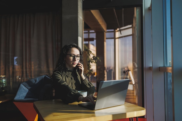 Jeune femme indépendante travaillant avec un ordinateur portable dans un espace de coworking et parlant au téléphone assis dans un espace ouvert avec une tasse de café et souriant tout en tenant des lunettes