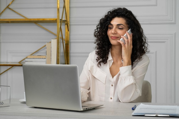 Jeune femme indienne aux cheveux bouclés parle sur un téléphone portable tout en travaillant avec un ordinateur portable à un bureau