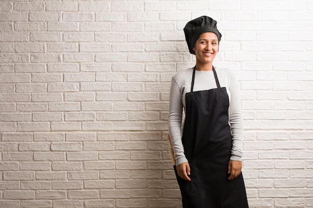 Jeune femme indienne de boulanger contre un mur de briques gaie et avec un grand sourire