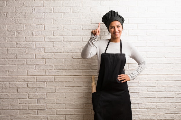 Jeune femme indienne de boulanger contre un mur de briques montrant le numéro un, symbole de comptage, concept de mathématiques, confiant et de bonne humeur