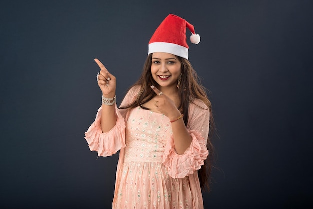 Jeune femme indienne célébrant Noël souriant et pointant quelque chose sur un espace vide sur fond gris concept de Noël