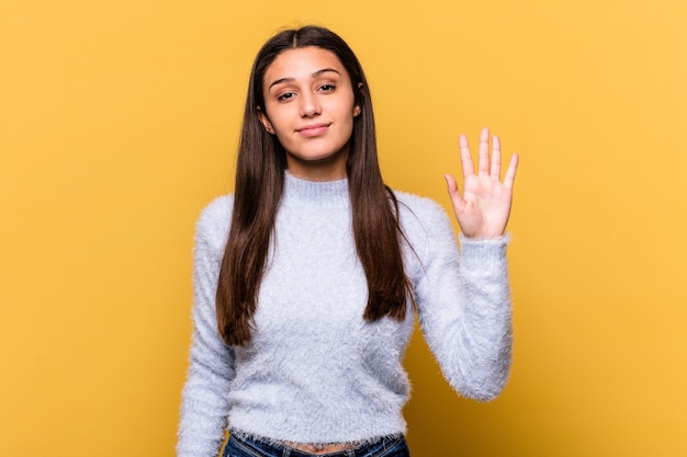 Jeune femme indienne isolée sur fond jaune souriant joyeux montrant le numéro cinq avec les doigts.