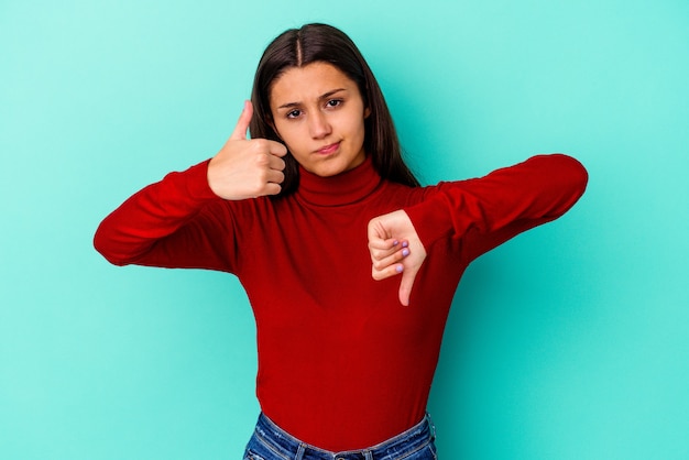 Jeune femme indienne isolée sur un mur bleu montrant les pouces vers le haut et les pouces vers le bas, difficile de choisir le concept