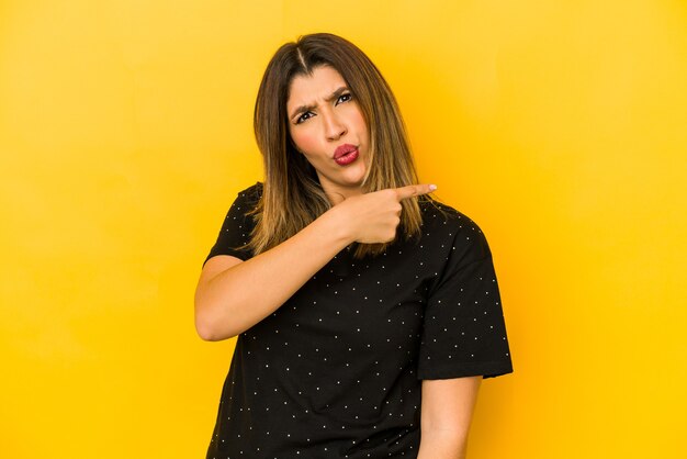 Jeune femme indienne isolée sur un mur jaune souriant et pointant de côté, montrant quelque chose à l'espace vide.