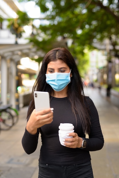 Jeune Femme Indienne Avec Masque à L'aide De Téléphone Et Prendre Un Café Sur Le Pouce Comme La Nouvelle Norme Pendant Le Covid-19 Dans La Ville