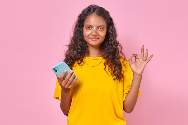 Une jeune femme indienne optimiste avec un téléphone montre un geste ok se tient dans le studio