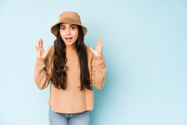 Jeune femme indienne portant un chapeau bleu recevant une agréable surprise, excitée et levant les mains.