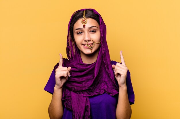 Jeune femme indienne portant un vêtement sari traditionnel isolé sur fond jaune indique avec les deux doigts avant montrant un espace vide.