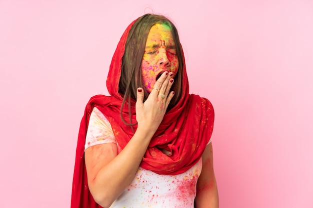 Jeune femme indienne avec des poudres de holi colorées sur son visage isolé sur le mur rose bâillant et couvrant la bouche grande ouverte avec la main