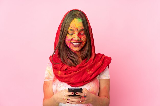 Jeune femme indienne avec des poudres de holi colorées sur son visage isolé sur un mur rose en envoyant un message avec le mobile