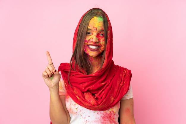 Jeune femme indienne avec des poudres de holi colorées sur son visage isolé sur un mur rose montrant et levant un doigt en signe du meilleur