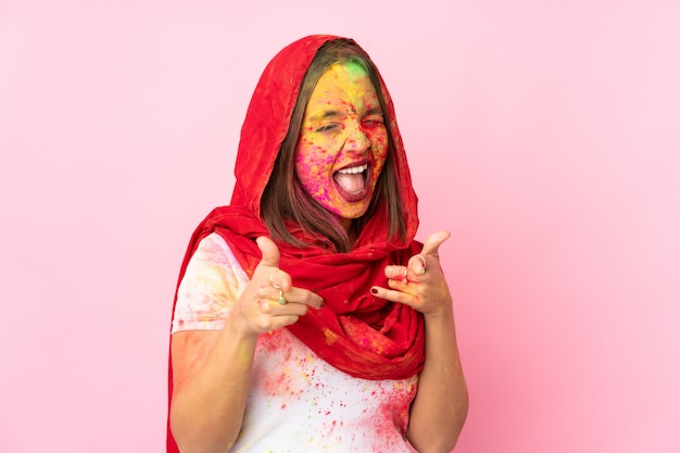 Jeune femme indienne avec des poudres de holi colorées sur son visage isolé sur un mur rose pointant vers l'avant et souriant