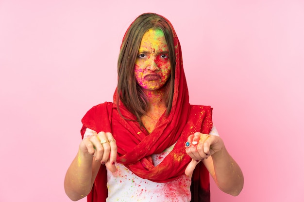 Jeune femme indienne avec des poudres de holi colorées sur son visage sur le mur rose montrant le pouce vers le bas avec deux mains