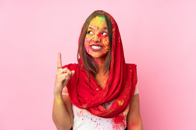 Jeune femme indienne avec des poudres de holi colorées sur son visage sur le mur rose pointant vers le haut une bonne idée