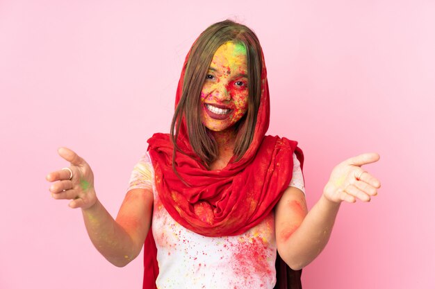 Jeune femme indienne avec des poudres de holi colorées sur son visage sur un mur rose présentant et invitant à venir avec la main