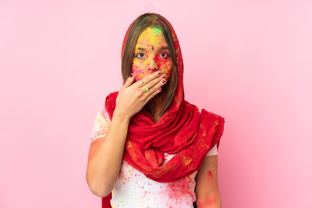 Jeune femme indienne avec des poudres de holi colorées sur son visage sur un revêtement mural rose bouche avec la main