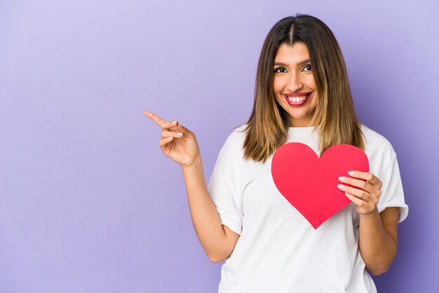 Jeune femme indienne tenant un coeur de Saint Valentin isolé souriant et pointant de côté, montrant quelque chose à l'espace vide.