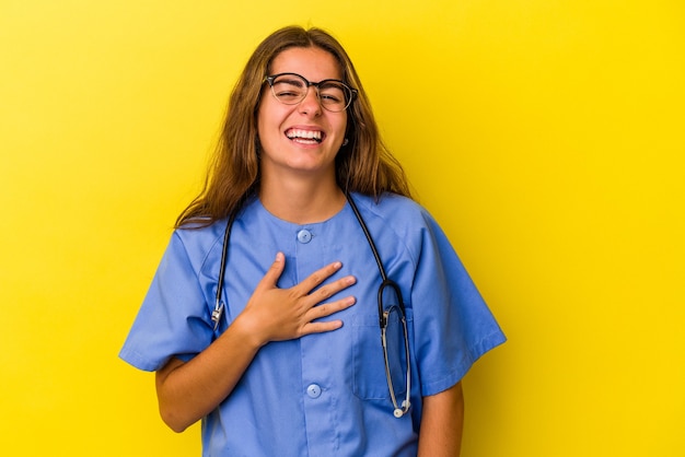 Jeune femme infirmière isolée sur fond jaune éclate de rire en gardant la main sur la poitrine.