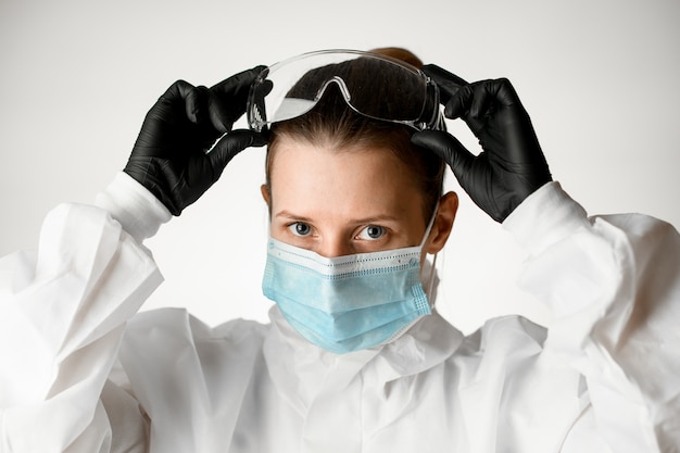 Jeune femme infirmière en masque médical et combinaison de protection blanche ajuste des lunettes sur sa tête