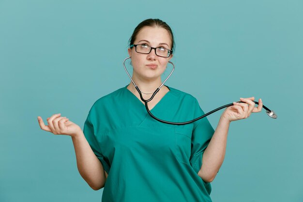 Jeune femme infirmière en uniforme médical avec stéthoscope autour du cou regardant confus répandre les bras sur les côtés debout sur fond bleu