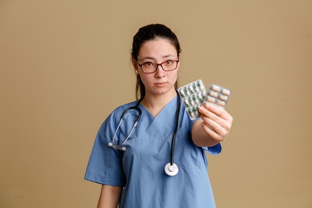 Jeune femme infirmière en uniforme médical avec stéthoscope autour du cou tenant des pilules montrant à la caméra avec un visage sérieux debout sur fond marron