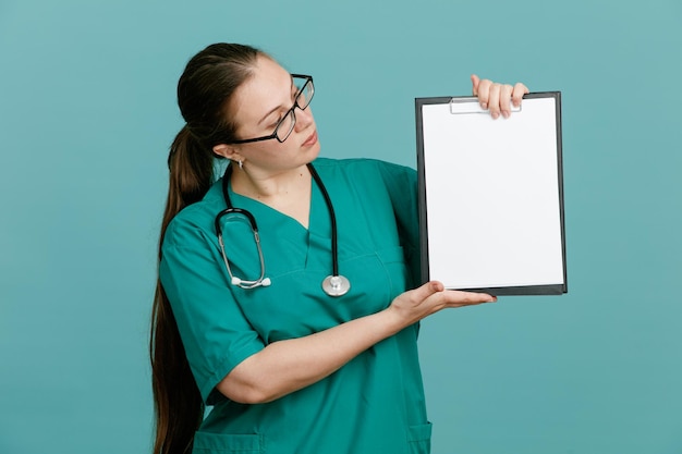 Jeune femme infirmière en uniforme médical avec stéthoscope autour du cou tenant le presse-papiers en le regardant avec une expression sérieuse confiante debout sur fond bleu