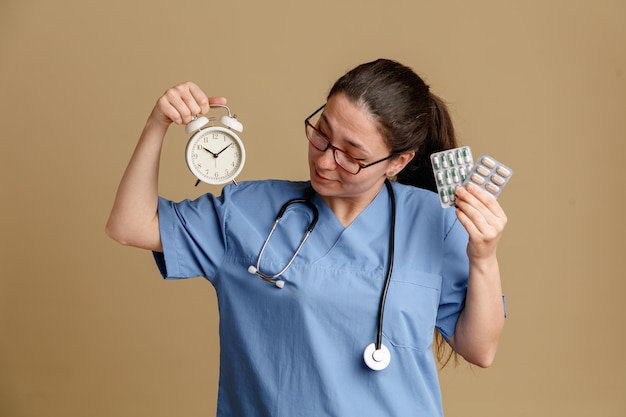 Jeune femme infirmière en uniforme médical avec stéthoscope autour du cou tenant un réveil et des pilules regardant l'horloge avec le sourire sur le visage debout sur fond marron