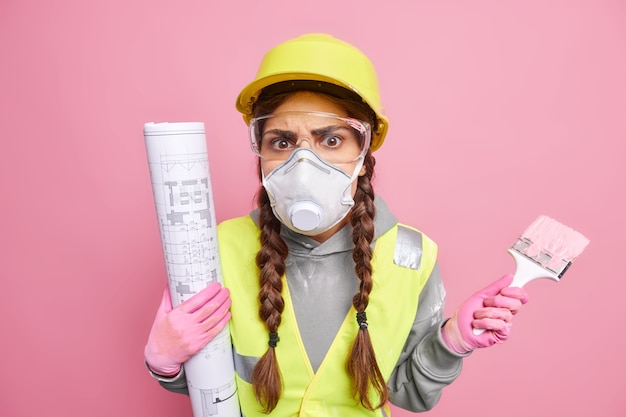 Une jeune femme ingénieure ou peintre sérieuse occupée à restaurer et à peindre des murs dans un nouvel appartement pose avec un plan et un pinceau