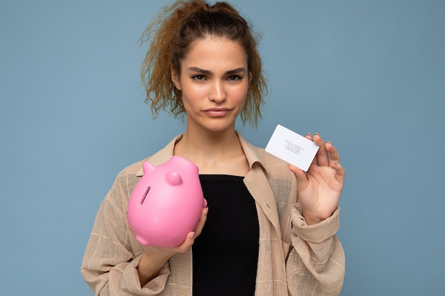 Jeune femme insatisfaite triste triste et séduisante brune bouclée avec des émotions sincères portant des vêtements élégants isolés sur fond bleu avec espace de copie, détient une tirelire rose et une carte de crédit pour