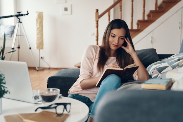 Jeune femme inspirée en vêtements décontractés écrivant quelque chose tout en passant du temps à la maison