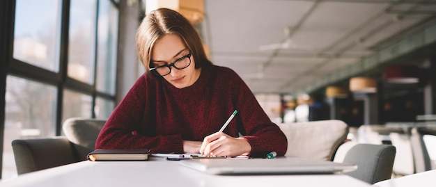 Jeune femme intelligente et intelligente à lunettes de 20 ans assise à table au café et écrivant