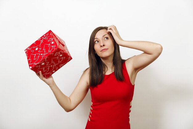 Une jeune femme intéressée et engagée vêtue d'une robe rouge et d'un chapeau de Noël tient une boîte-cadeau, pense qu'elle a été donnée sur fond blanc. Fille de Santa avec présent isolé. Concept de vacances de nouvel an 2018