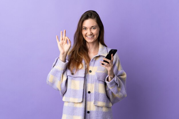 Jeune femme d'Irlande isolée sur fond violet à l'aide de téléphone mobile et faisant signe OK