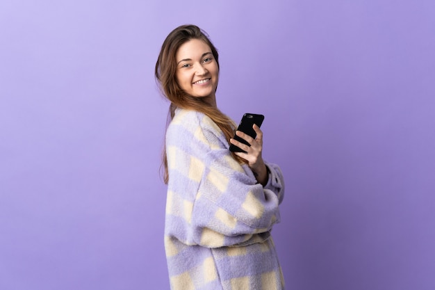 Jeune femme d'Irlande isolée sur fond violet tenant un téléphone mobile et les bras croisés