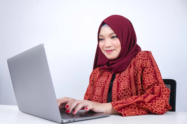 Une jeune femme islamique asiatique est assise et sourit en travaillant sur un ordinateur portable sur fond blanc